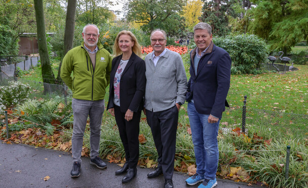 Vier Personen lächeln freundlich in einem Park mit buntem Herbstlaub, umgeben von grüner Vegetation.